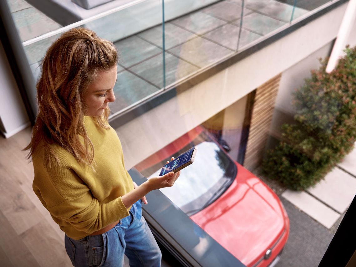 Ženska gleda v mobilni telefon. V ozadju je rdeči Volkswagen. V zgornjem desnem kotu sta logotipa trgovin App Store in Google Play.