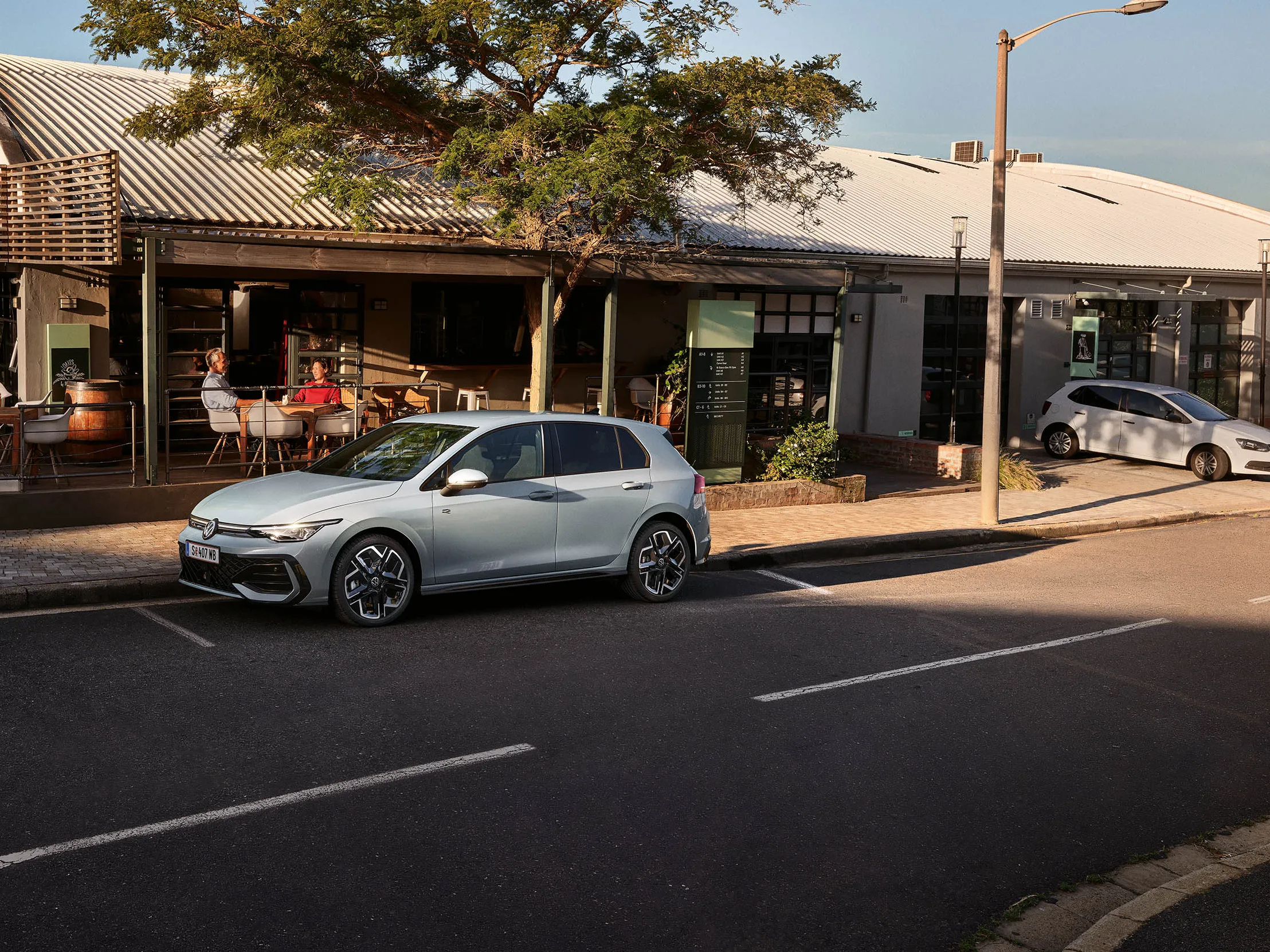 Pogled od spredaj na bel VW Golf eHybrid s priključenim polnilnim kablom, dva moška se približujeta vozilu