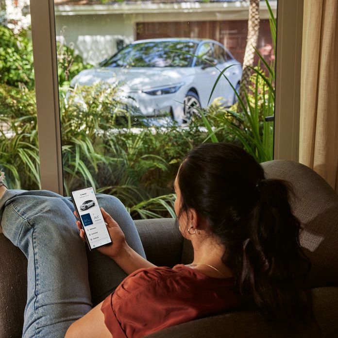 Ženska s pametnim telefonom v roki sproščeno sedi v dnevni sobi in skozi okno gleda VW ID.7.