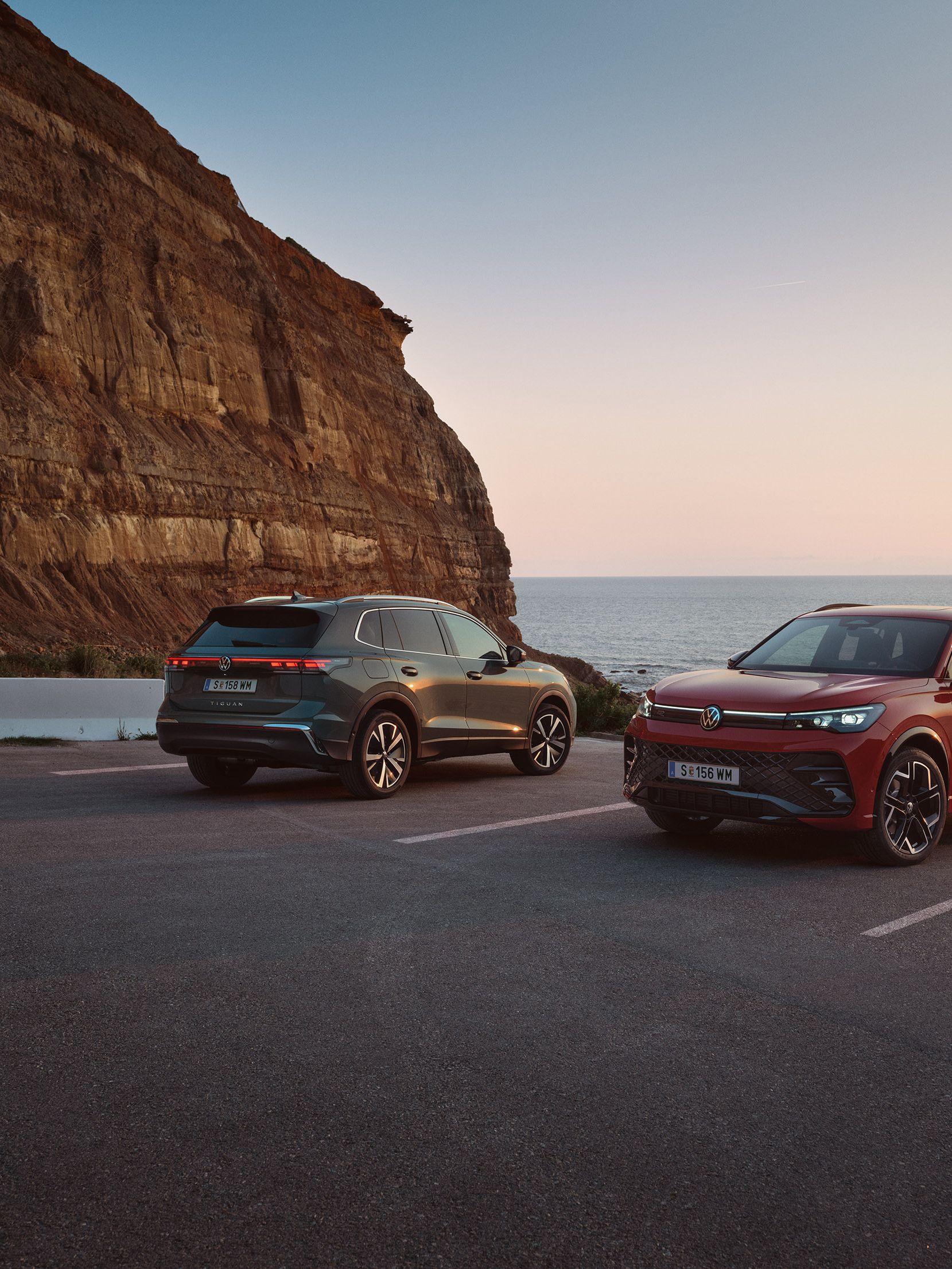 Dva VW Tiguan v temnozeleni in rdeči barvi sta parkirana pred skalnato obalno pokrajino.