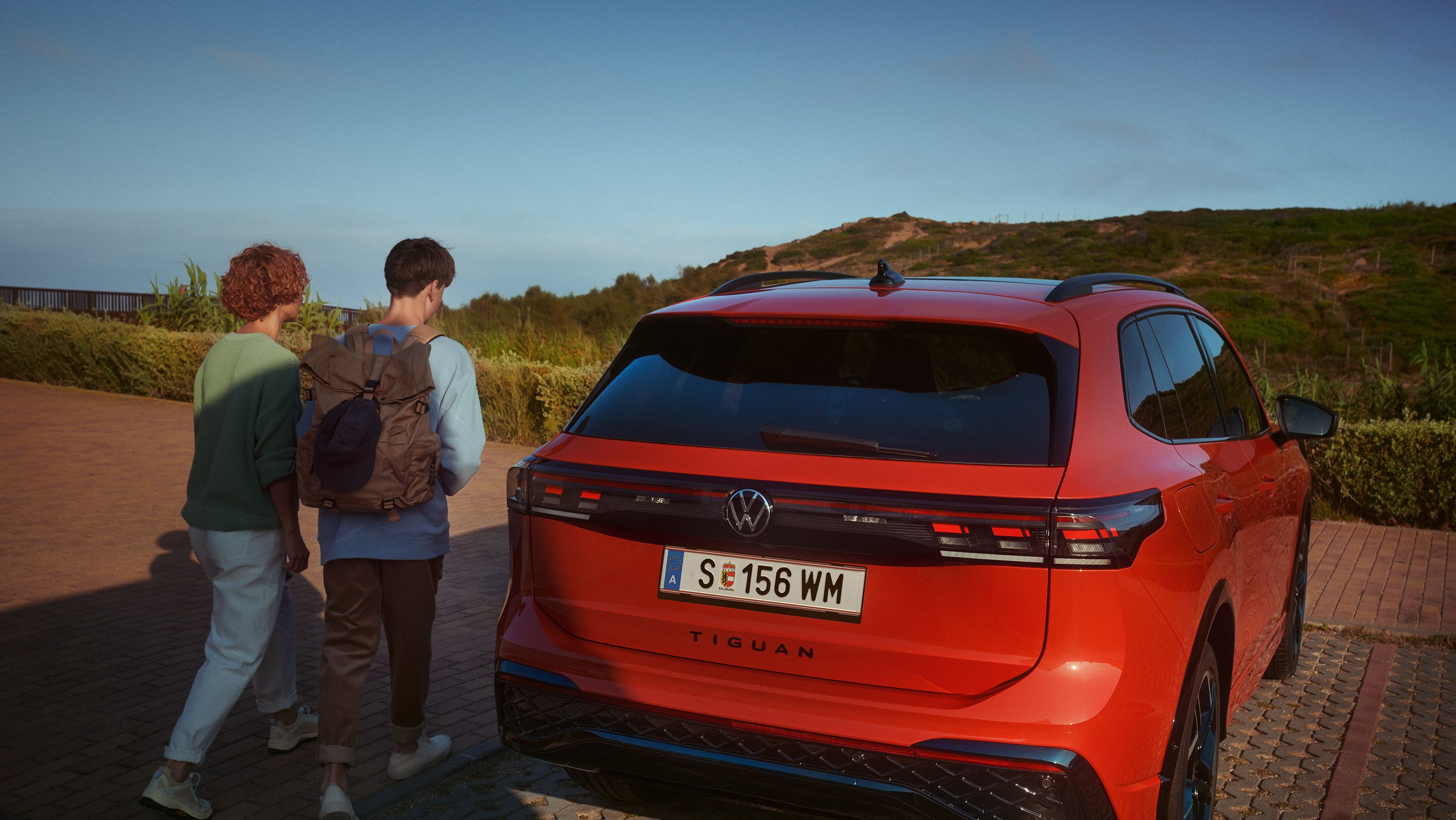 Pogled na zadek parkiranega VW Tiguana. Avtomobilu se približujeta ženska in najstnik. 