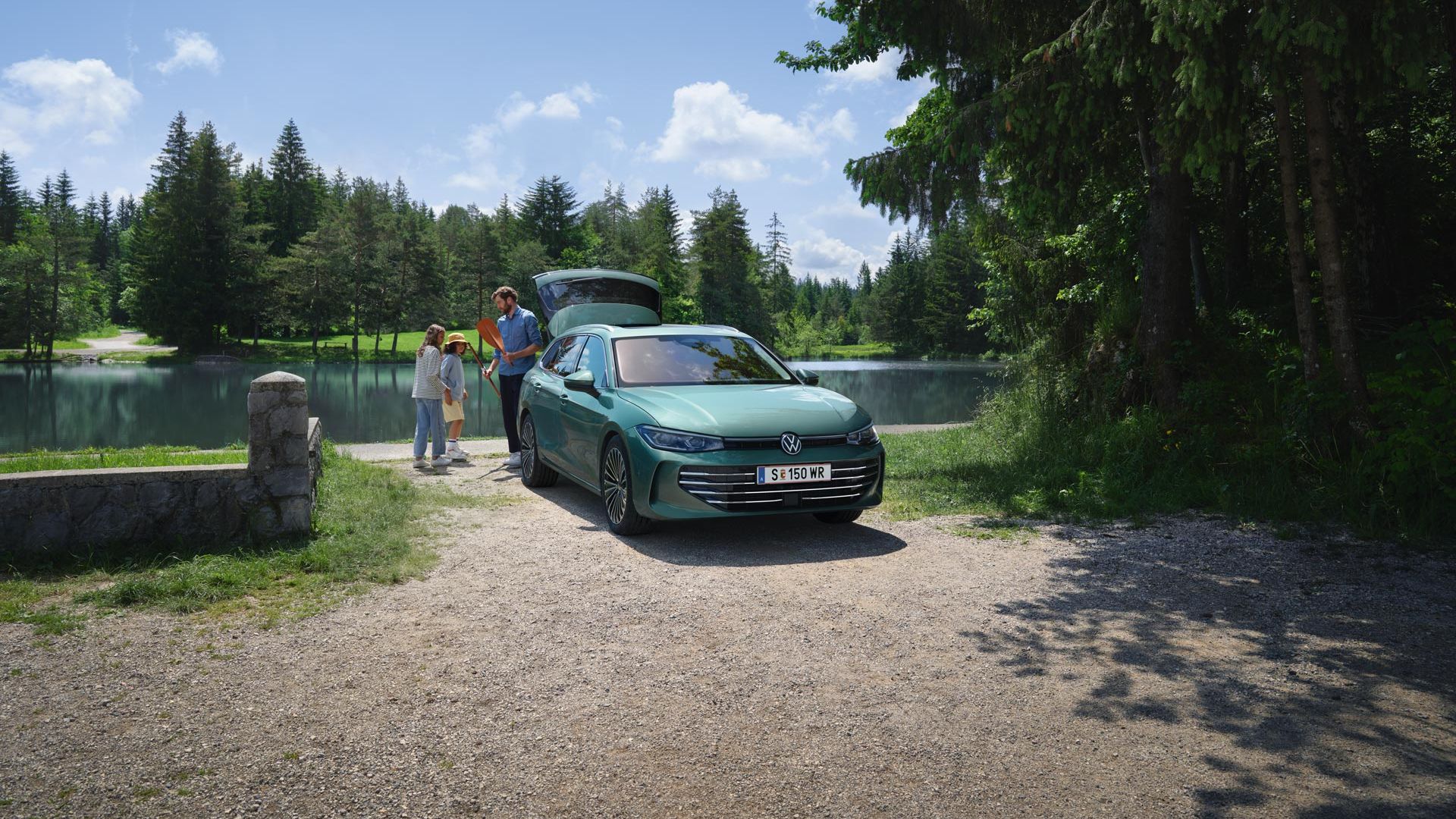 VW Passat in blaugrün parkt an einem Badesee mit geöffnetem Kofferraum. Ein Mann verteilt Freitzeitspielzeug an zwei Kinder.