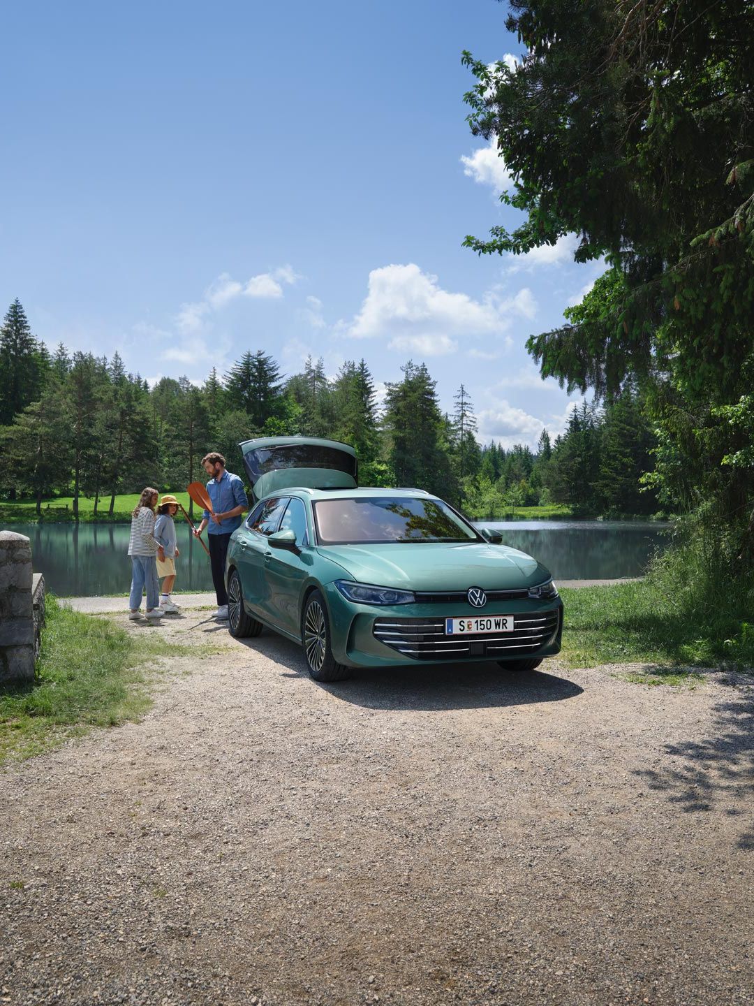 VW Passat in blaugrün parkt an einem Badesee mit geöffnetem Kofferraum. Ein Mann verteilt Freitzeitspielzeug an zwei Kinder.