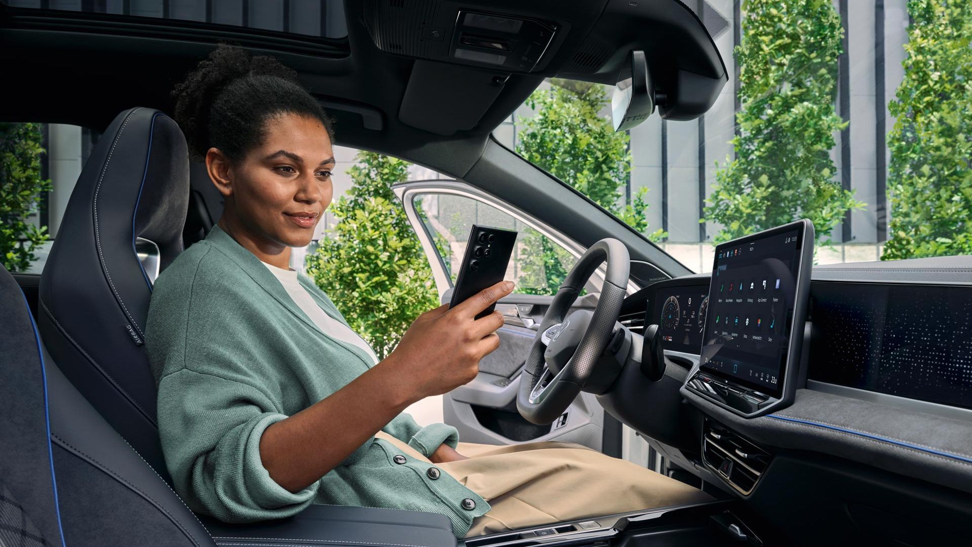 Eine Frau sitzt bei geöffneter Tür auf dem Fahrersitz des geparkten VW Passat und blickt auf ihr Smartphone.