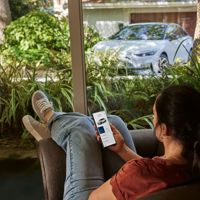 Ženska s pametnim telefonom v roki sproščeno sedi v dnevni sobi in skozi okno gleda VW ID.7.