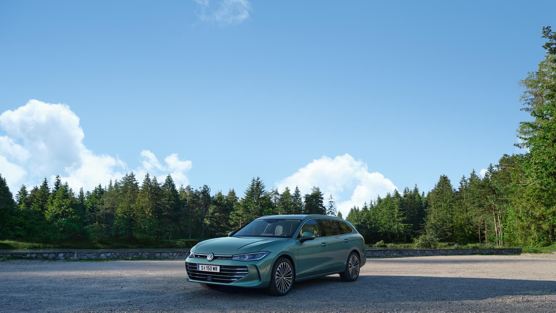 VW Passat in blaugrün steht auf einer Kiesfläche, das Gelände grenzt an einen Tannenwald.