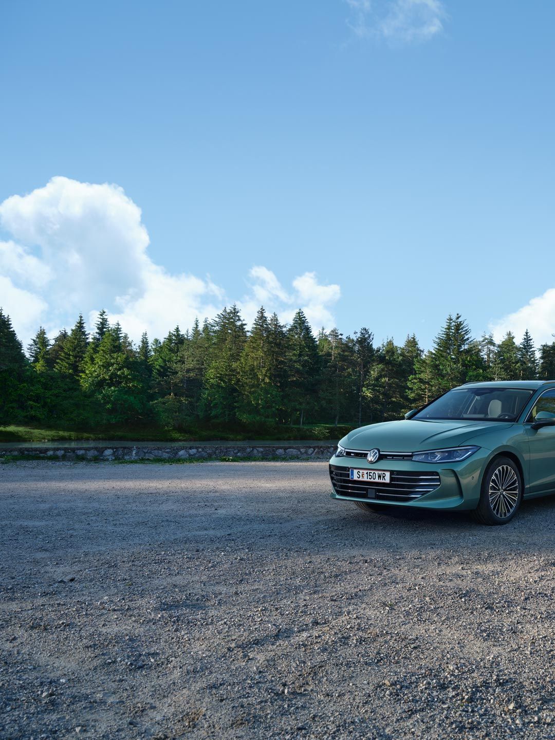 VW Passat in blaugrün steht auf einer Kiesfläche, das Gelände grenzt an einen Tannenwald.